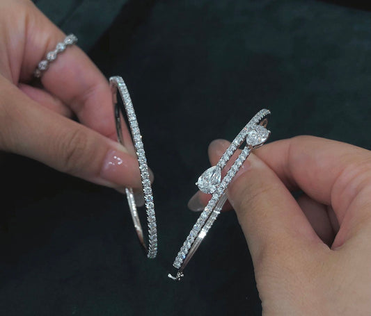 Two silver bracelets with diamond accents held by hands against a dark background